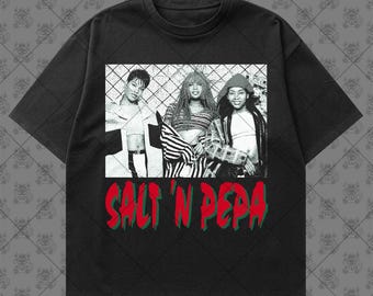 May include: A black short-sleeve t-shirt featuring a black and white photographic print of the musical group Salt-N-Pepa in front of a chain-link fence. Below the image, the text SALT 'N PEPA is printed in bold red letters with green outlines.