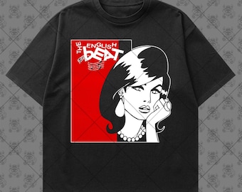 May include: A black short-sleeve t-shirt featuring a graphic print of a woman with dark hair and a pearl necklace. The design includes a red square background, musical notes, and the white text "THE ENGLISH BEAT" in a bold, stylized font.