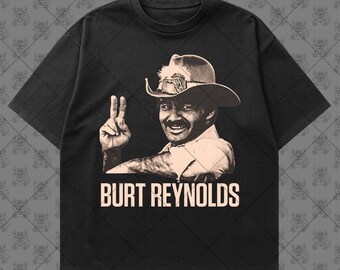 May include: A black short-sleeve t-shirt featuring a light-colored graphic print of Burt Reynolds wearing a cowboy hat and making a peace sign gesture. The text "BURT REYNOLDS" is printed in a bold, light-colored font directly below the portrait.