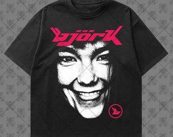 May include: A black short-sleeve t-shirt featuring a high-contrast white portrait of Björk on the front. The word "björk" is printed in a bold, stylized pink font above the face, with a small pink circular logo located below the chin area.
