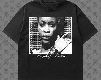 May include: A black short-sleeve t-shirt featuring a central white graphic print of Erykah Badu. The design includes a repeating 'BADUIZM' text pattern in the background and the name 'Erykah Badu' printed in a cursive script below the portrait.