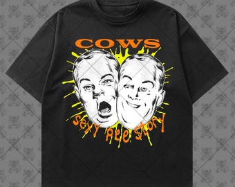 May include: A black short-sleeve t-shirt featuring a graphic print of two faces in white line art. The text COWS appears in orange above the faces, and the text sexy pee story appears in orange below the faces, all set against a yellow splatter.