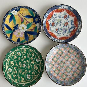 Puede incluir: Cuatro platos redondos de cerámica con bordes festoneados, cada uno con patrones distintos: uno con motivos florales amarillos, uno con diseños florales geométricos rojos y azules, uno con enredaderas verdes y uno con un patrón de celosía azul y naranja.