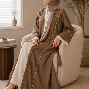 Könnte beinhalten: Ein langärmeliges, bodenlanges braunes Abaya mit einer cremefarbenen Innenschicht, aufwendiger Spitzenborte entlang der Vorderöffnung und passendem braunen Hijab. Das Kleidungsstück verfügt über weiße Paspeldetails an den Ärmeln und eine Taillenbindung.