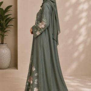 Puede incluir: Una abaya de color verde salvia con mangas largas y un hijab a juego. La prenda está adornada con delicados bordados florales en tonos rosa suave y blanco a lo largo de las mangas y la parte inferior de la falda, creando un aspecto coherente.