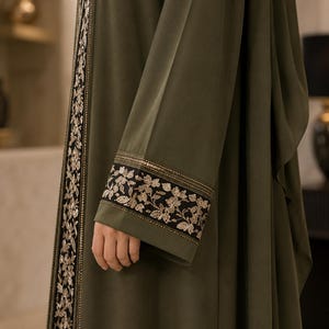 Puede incluir: Una abaya de color verde oliva con mangas largas y puños negros adornados con intrincados bordados florales dorados. La abertura frontal de la prenda está detallada con un panel negro a juego que presenta el mismo patrón floral dorado.