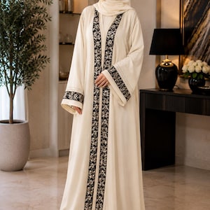 May include: A floor-length cream-coloured abaya featuring a long, vertical black panel down the centre and on the sleeve cuffs. The black panels are decorated with a detailed silver and white floral embroidery pattern. The garment includes a matching cream-coloured hijab that covers the head and neck, creating a cohesive and layered appearance.