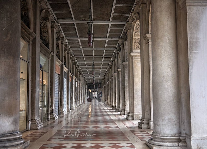 May include: A long, narrow, arched hallway with a tiled floor and a white ceiling. The hallway is lined with columns on either side.