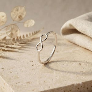 Könnte beinhalten: Ein polierter Silberring mit einem zarten Unendlichkeitssymbol-Design. Der Ring ist dünn und kreisförmig und weist eine glatte, reflektierende Metalloberfläche auf, die das Licht einfängt. Die Unendlichkeitsschleife ist oben auf dem Ring zentriert.