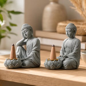 May include: Two grey stone-textured Buddha figurines, each seated on a lotus-shaped base. One Buddha rests a hand on its cheek, while the other sits in a meditative pose. Each figure features a small, conical incense cone placed in a gold ring.