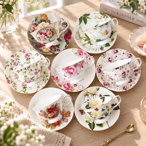 Op de afbeelding: Een verzameling van zes porseleinen theekopjes en schoteltjes op een houten ondergrond. Elk item heeft unieke bloemmotieven, waaronder rozen, witte bloesems en kleurrijke boeketten, afgewerkt met goudkleurige randen en sierlijke oren.