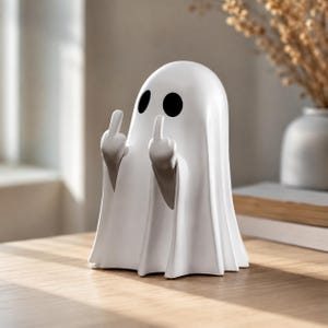 Peut inclure: Une figurine de fantôme blanche avec une finition lisse et mate, dotée de deux grands yeux noirs circulaires. Le fantôme est représenté avec les deux bras levés, les majeurs tendus dans un geste, avec un corps drapé en forme de drap.