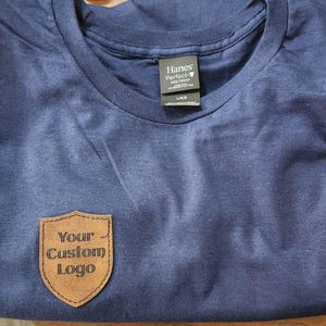 May include: A navy blue crew neck t-shirt featuring a brown leather-like shield-shaped patch on the lower left side. The patch is embossed with the text "Your Custom Logo" in a dark font. The shirt has a Hanes Perfect-T label at the neckline.