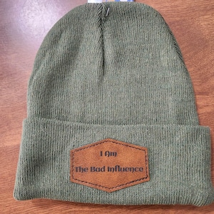 Può includere: Un berretto in maglia verde oliva con risvolto. Al centro del risvolto è applicata una toppa esagonale in similpelle marrone con il testo "I Am The Bad Influence" impresso in un carattere marrone scuro senza grazie su due righe.