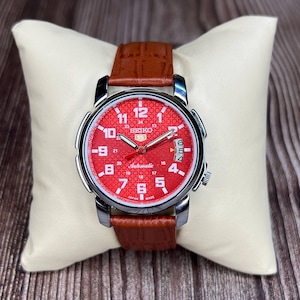 Op de afbeelding: Een Seiko 5 automatisch polshorloge met een levendige rode textuurwijzerplaat met witte Arabische cijfers, zilverkleurige wijzers en een datumvenster op de drie-uurpositie. Het horloge heeft een gepolijste zilverkleurige metalen kast en een bruine leren band.