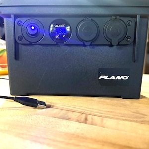 Puede incluir: Una caja de almacenamiento Plano de color gris oscuro con un panel de control frontal que incluye una pantalla digital de voltaje iluminada en azul, un puerto de alimentación circular y dos tomas cubiertas etiquetadas como USB y 12V. Un cable USB negro está en primer plano.