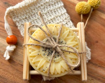 May include: A round, pale yellow sunflower-shaped soap bar tied with twine, resting on a small wooden rack. Behind it sits a cream-coloured crocheted soap pouch with an orange bead, accompanied by two small, round, yellow dried flower heads.