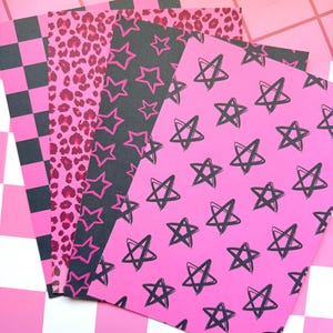 May include: Four rectangular sheets of paper featuring distinct patterns. One sheet has a black and pink checkerboard print, one has a pink and dark pink leopard print, one is black with pink stars, and one is pink with hand-drawn black stars.