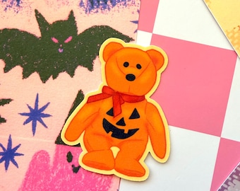 May include: An orange teddy bear sticker featuring a black jack-o'-lantern face on its chest and a red ribbon tied around its neck. The bear has black eyes and a black nose, with a yellow border outlining the entire shape of the sticker.