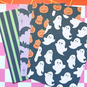 May include: Four rectangular sheets of patterned paper featuring Halloween themes. One sheet has green and black vertical stripes, another displays purple bats, a third shows orange jack-o'-lanterns on black, and the fourth features white ghosts on black.