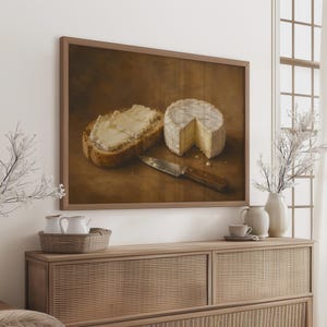 Op de afbeelding: Een ingelijste muurkunstprint met een stilleven van een snee brood met boter, een ronde kaas met een weggesneden punt en een metalen mes met een houten handvat, alles tegen een donkerbruine achtergrond.