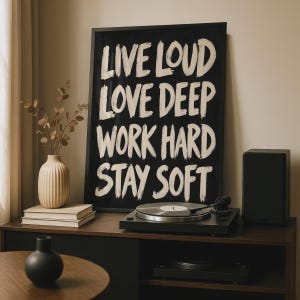 Könnte beinhalten: Ein rechteckiges, schwarz gerahmtes Wandbild mit weißer Pinselstrich-Schrift, die LIVE LOUD, LOVE DEEP, WORK HARD, STAY SOFT lautet. Der Text ist in vier Zeilen auf einem einfarbigen schwarzen Hintergrund innerhalb des Rahmens angeordnet.