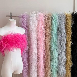 Pode incluir: Um top sem alças coberto de penas rosa brilhante exibido num manequim, ao lado de um suporte metálico com uma coleção de longas boas de penas em várias cores, incluindo lavanda, branco, rosa, bege, verde menta, cinzento, dourado e preto.