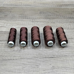 Peut inclure: Cinq bobines cylindriques de fil à coudre marron foncé disposées en rangée horizontale. Chaque bobine présente un noyau en plastique blanc visible aux deux extrémités, avec le fil enroulé serré autour du centre de manière uniforme.