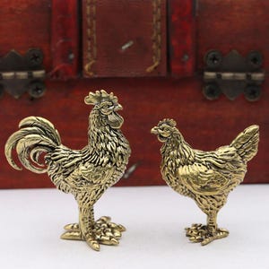 May include: Two small brass figurines depicting a rooster and a hen. The rooster stands on the left with detailed plumage and a prominent comb, while the smaller hen stands on the right. Both figures feature a metallic gold finish with dark patina.