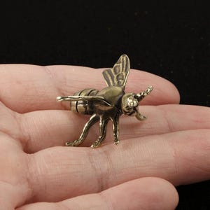 May include: A small, three-dimensional bee figurine crafted from a metallic material with an antique brass finish. The insect features detailed wings, a segmented abdomen, and six legs, showcasing a textured surface with dark patina accents.