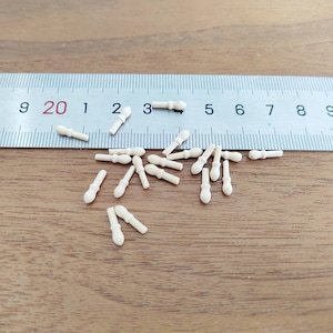 May include: A collection of small, off-white, solid-colored miniature pegs or spindles scattered below a metal ruler. Each piece features a rounded head and a straight, cylindrical body. The ruler shows these items measure approximately 0.5 inches.