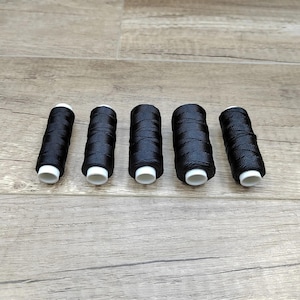May include: Five cylindrical spools of black sewing thread arranged in a horizontal row. Each spool features a white plastic core visible at both ends, with the black thread tightly wound around the center of the cylindrical structure.