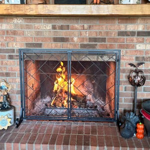 Puede incluir: Una pantalla de chimenea de metal negro con un diseño de dos paneles con bisagras y un patrón de malla de celosía diagonal. La pantalla se sitúa frente a una chimenea de ladrillo con un fuego de leña de llamas naranjas y amarillas.