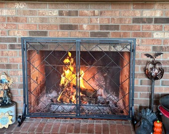 May include: A black metal fireplace screen with a two-panel hinged design and a diagonal lattice mesh pattern. The screen stands in front of a brick fireplace hearth containing a burning wood fire with bright orange and yellow flames.