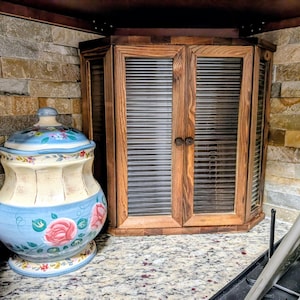 May include: A wooden corner cabinet with two doors featuring horizontal ribbed glass panels and small round knobs, positioned next to a ceramic canister decorated with hand-painted pink roses and blue floral accents on a cream background.