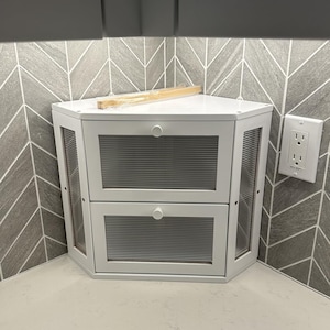 May include: A white corner storage unit featuring two drawers with horizontal ribbed glass panels and small round knobs. The unit has angled side panels with matching ribbed glass inserts and a flat top surface with a thin wooden strip resting on it.