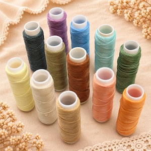 May include: A collection of twelve spools of waxed thread in various colors, including shades of yellow, beige, brown, orange, green, blue, and purple. Each spool features a white plastic core and is arranged in a cluster on a textured fabric surface.