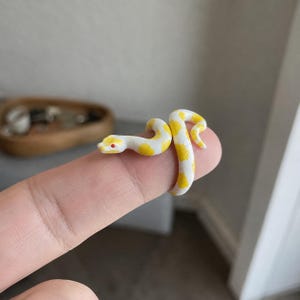 May include: A miniature white snake figurine with bright yellow spots and small red eyes. The snake is coiled in a curved shape, featuring a smooth, textured surface that mimics scales, designed to wrap around a finger as a decorative accessory.