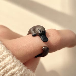 May include: An adjustable ring featuring a sculpted brown platypus with a dark black bill. The design wraps around the finger, with the platypus head positioned at one end and the tail at the other, creating a continuous animal-shaped band.