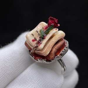 Può includere: Un anello in argento con una pila in miniatura di tre libri con copertine color crema e marrone. Una piccola e dettagliata rosa rossa con foglie verdi poggia sopra i libri. La base dell'anello presenta un bordo metallico decorato e smerlato.