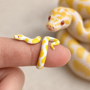 May include: A wrap-around ring shaped like a snake with a white and yellow spotted pattern and small red eyes. The ring is coiled around a finger, mimicking the appearance of a larger albino python snake visible in the background of the image.
