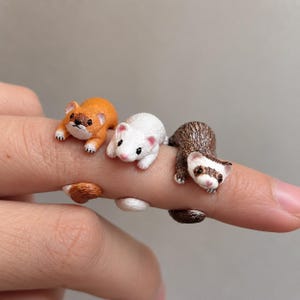 May include: Three adjustable rings featuring miniature ferret figures in orange, white, and brown. Each ring is designed with the ferret's body wrapping around the finger, showcasing detailed facial features, small ears, and a textured tail.