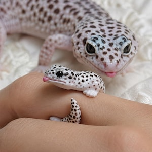 May include: A ring shaped like a small white leopard gecko with black spots and a tiny pink tongue sticking out. The gecko's body wraps around the finger, with its head resting on top and its tail curving underneath to complete the band design.