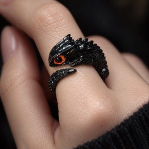 Può includere: Un anello nero a forma di piccola lucertola o drago con una superficie ruvida e squamosa. La creatura ha un unico occhio prominente di colore arancione brillante. L'anello è di tipo aperto, con la testa e la coda che si incontrano sul davanti.