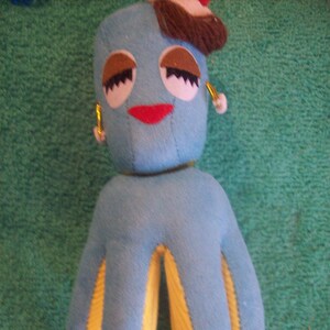 May include: A handmade blue plush octopus doll with yellow accents on its tentacles. The doll has brown eyelashes, a red mouth, and a small brown hat with a red and white detail. It also has gold earrings. The doll is on a green background.