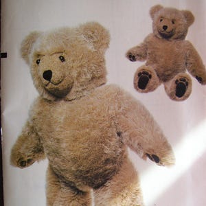 May include: A Vogue Patterns sewing pattern for a teddy bear craft. The pattern features a large, plush teddy bear with a light brown fur-like texture. The bear has a smiling face, black eyes, and a black nose. A smaller version of the bear is also shown.