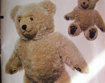 May include: A Vogue Patterns sewing pattern for a teddy bear craft. The pattern features a large, plush teddy bear with a light brown fur-like texture. The bear has a smiling face, black eyes, and a black nose. A smaller version of the bear is also shown.