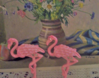 May include: Two pink plastic flamingo figurines with rhinestones on a white crocheted doily.