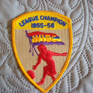 May include: Embroidered patch with a shield shape, featuring the text "LEAGUE CHAMPION 1955-56" in blue and yellow. A red figure is depicted playing with a ball, with a flag that reads "WABC". The patch has a yellow border.
