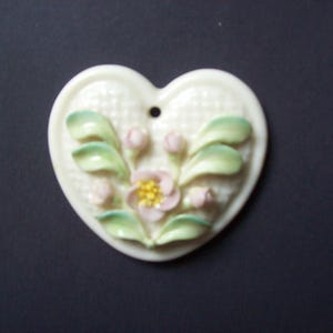 May include: Cream-colored heart-shaped ceramic ornament with a floral design. The ornament features a raised relief of a flower with pink petals and a yellow center, surrounded by green leaves and buds. A small hole is at the top.
